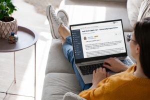 A college student lounges on a couch using a laptop that displays a course community page for “Fall 2026 UNIV 101 – University Experience,” with a welcome post from the instructor.